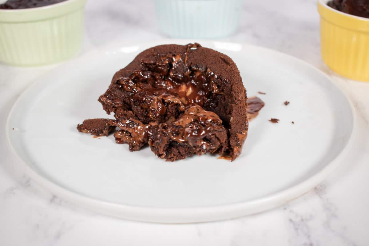 Vegan chocolate lava cake on a white plate. Chocolate drizzling out. Multi-coloured ramekins behind.