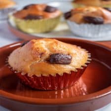 A vegan banana muffin inside a large brown ramekin. More muffins in the background inside their metallic cake cases.