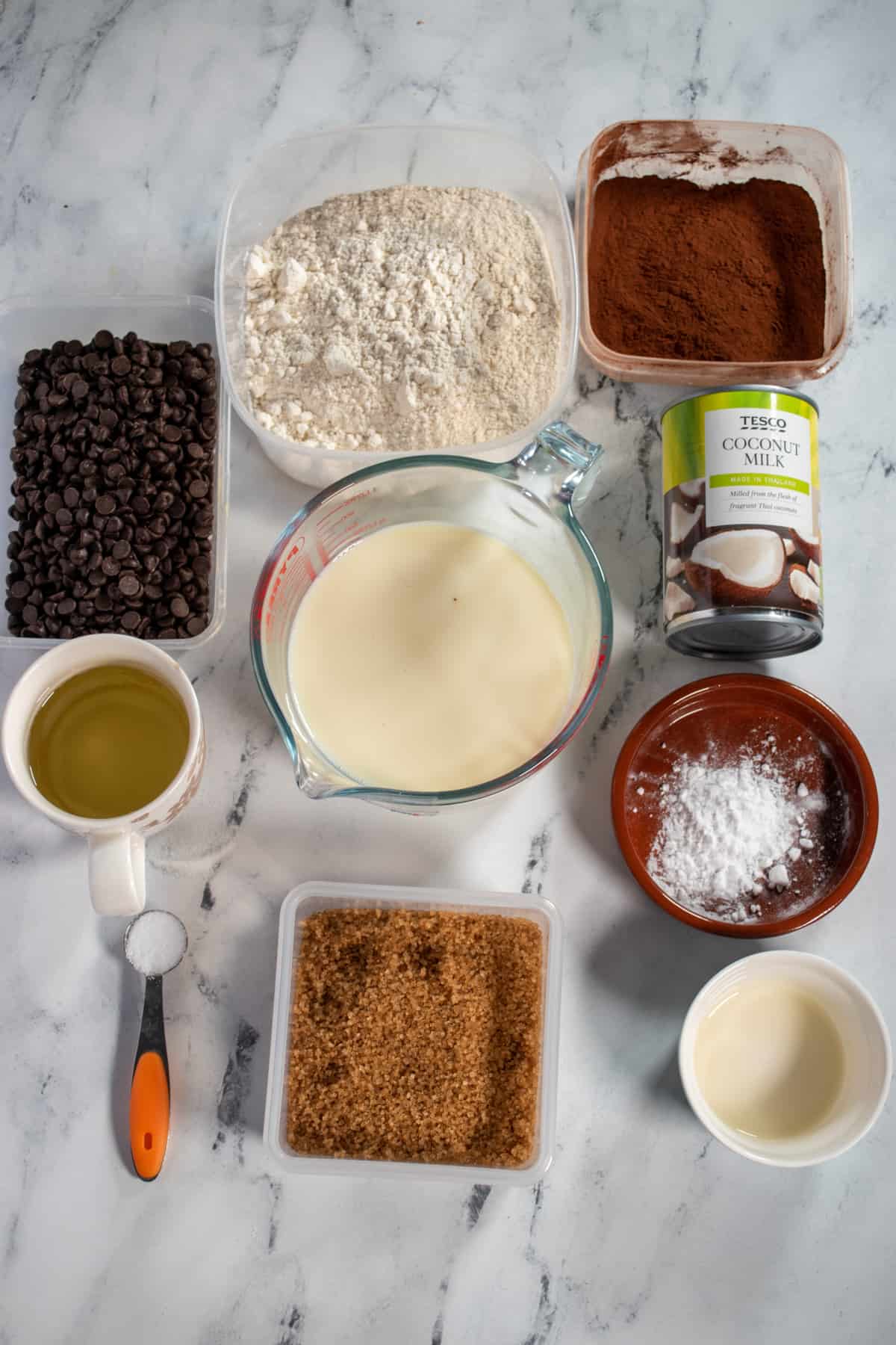 An aerial shot of the ingredients needed to make a vegan chocolate ganache cake. Flour, cocoa powder, plant milk, brown sugar, baking soda, salt, apple cider vinegar, vegetable oil, chocolate chips, coconut milk and powdered sugar.