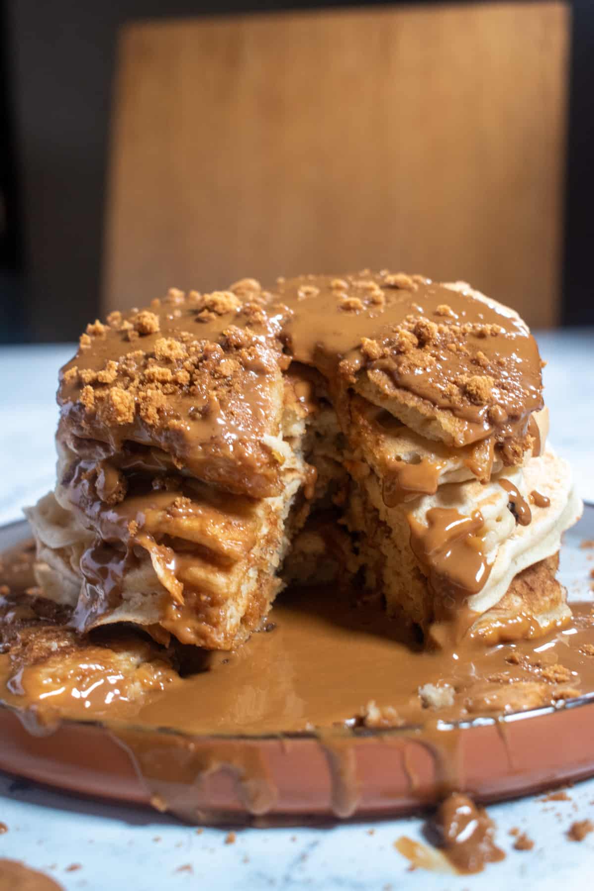 A large stack of vegan Biscoff pancakes on a brown plate. They're sat in a pool of Biscoff spread. There is a slice missing from the center of the stack.