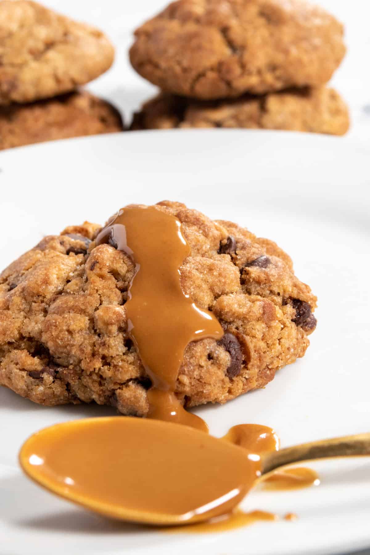 A chunky Biscoff cookie with melted biscoff spread on top of it. A golden spoon also with biscoff spread inside it. 