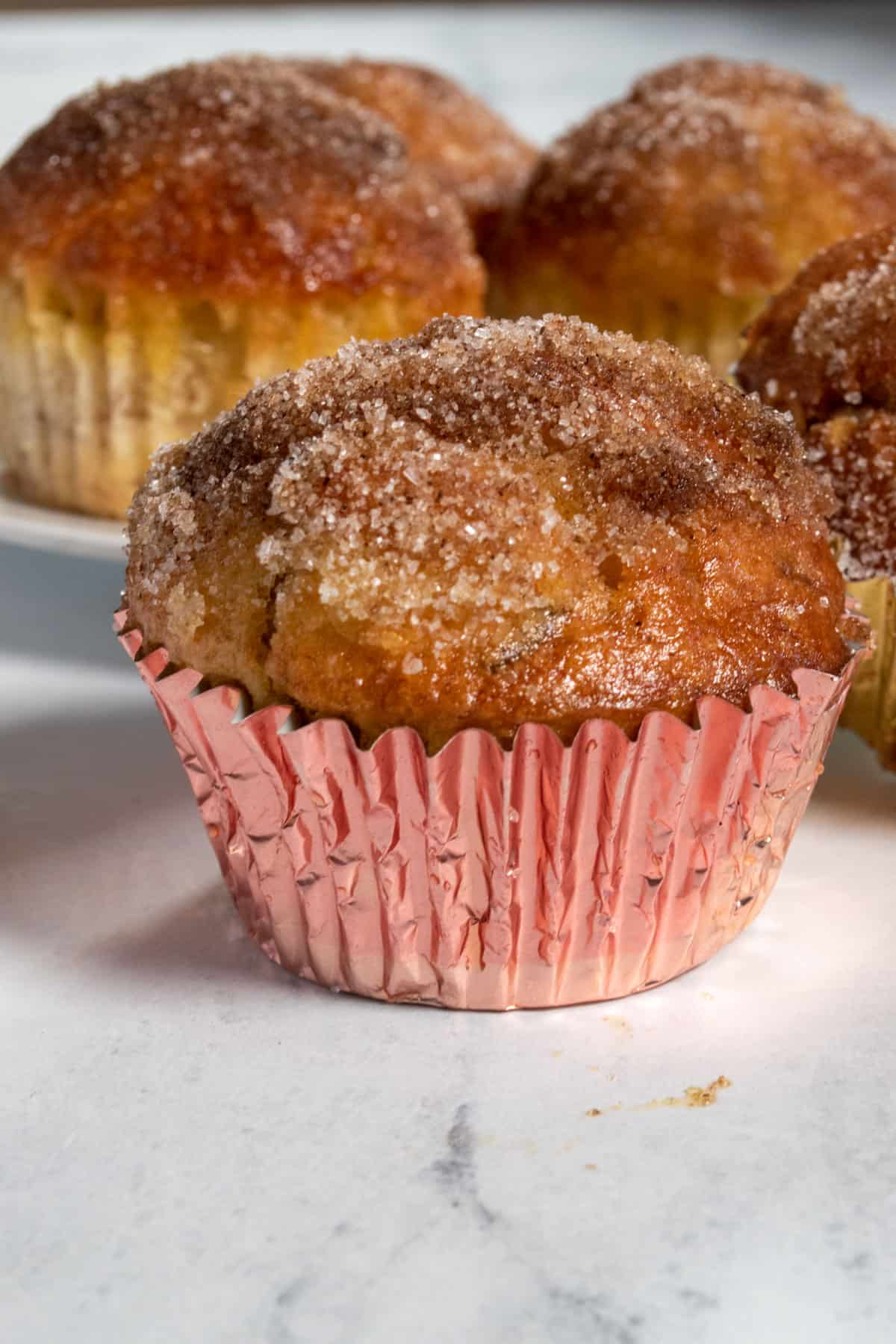 One of my golden brown vegan cinnamon muffins in a metallic pink liner. Several other muffins are sitting behind it.