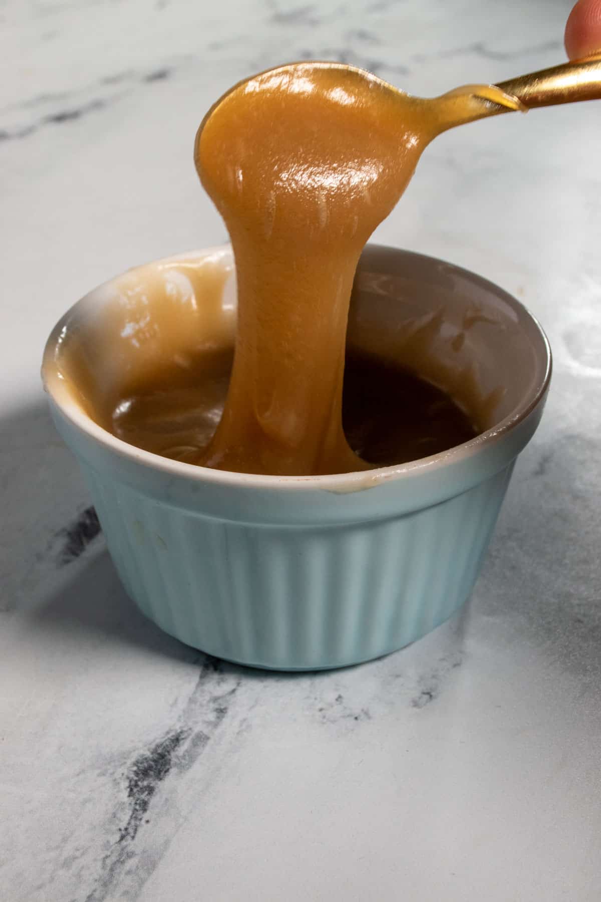 A blue ramekin filled with vegan dulce de leche. A golden spoon is drizzling it downards from above.