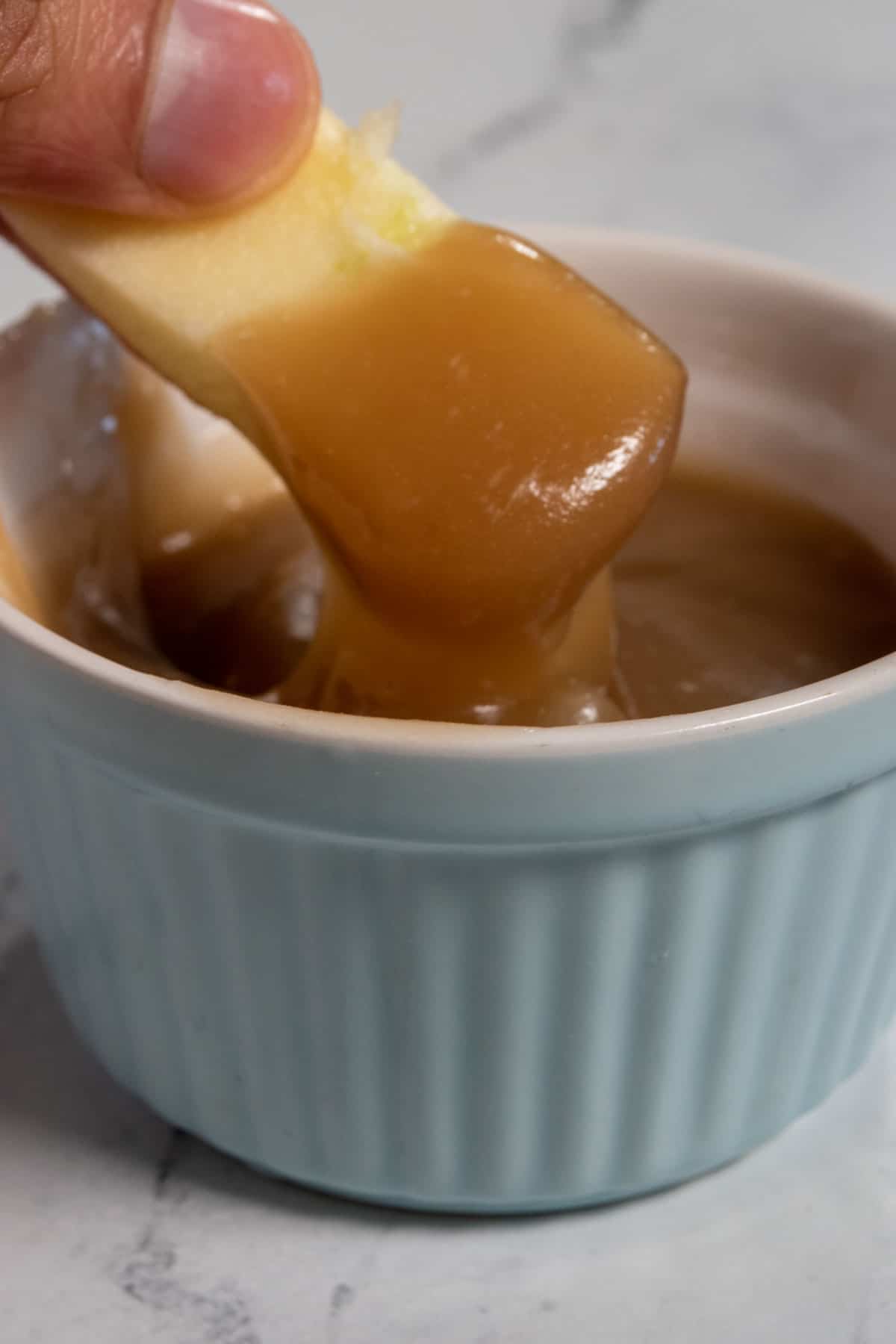 An apple piece being dipped into a blue ramekin full of vegan dulce de leche. 