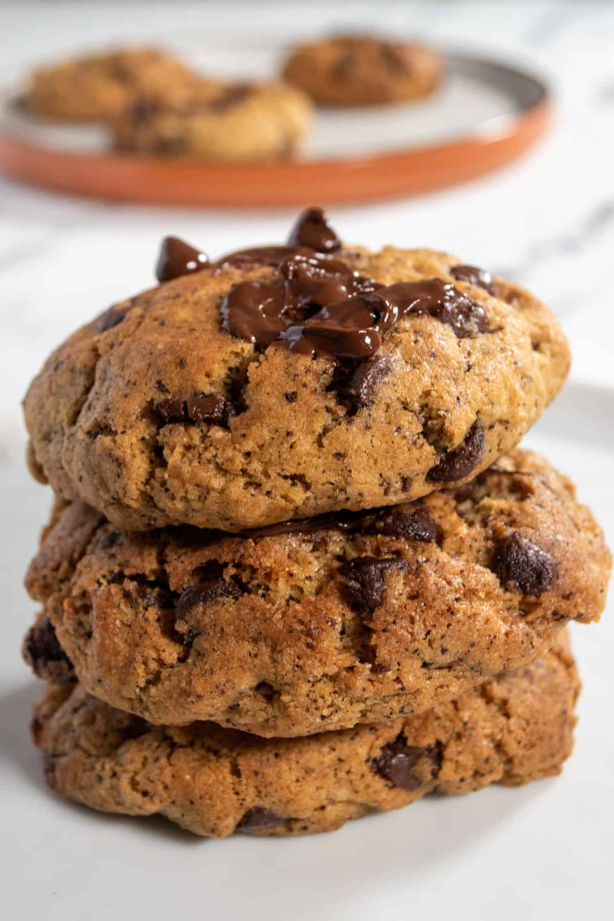 Three espresso chocolate chip cookies stacked on top of each other. Several others sit on a brown plate behind them.