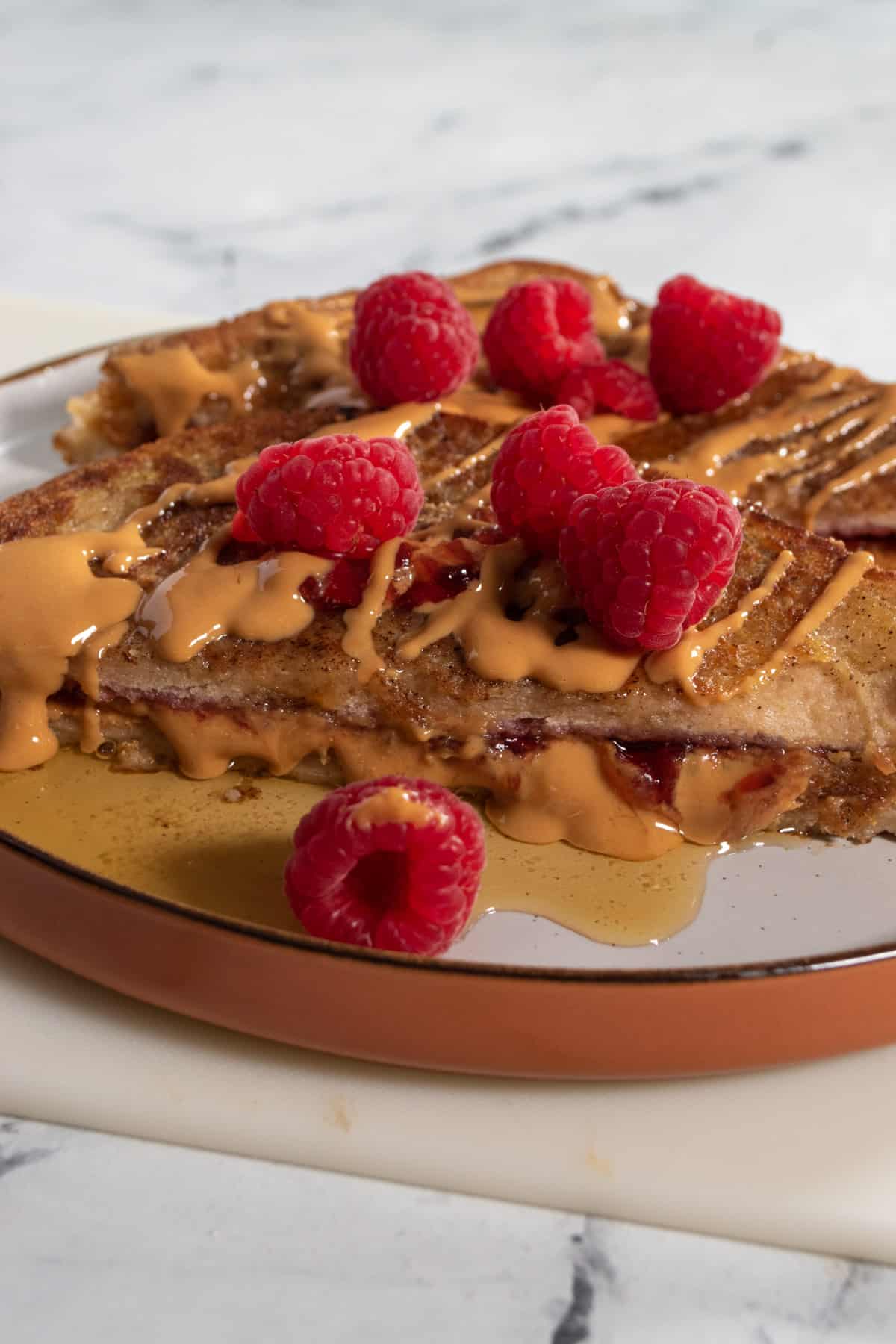 Vegan peanut butter and jelly French toast on a brown plate. Garnished with raspberries, maple syrup and peanut butter drizzle.