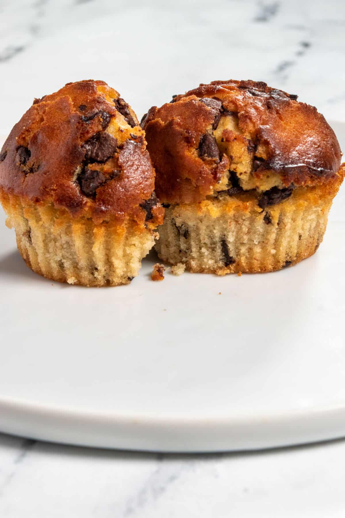 Two peanut butter muffins on a white plate. The chocolate chips stand out. One of the muffins is sliced in half. 