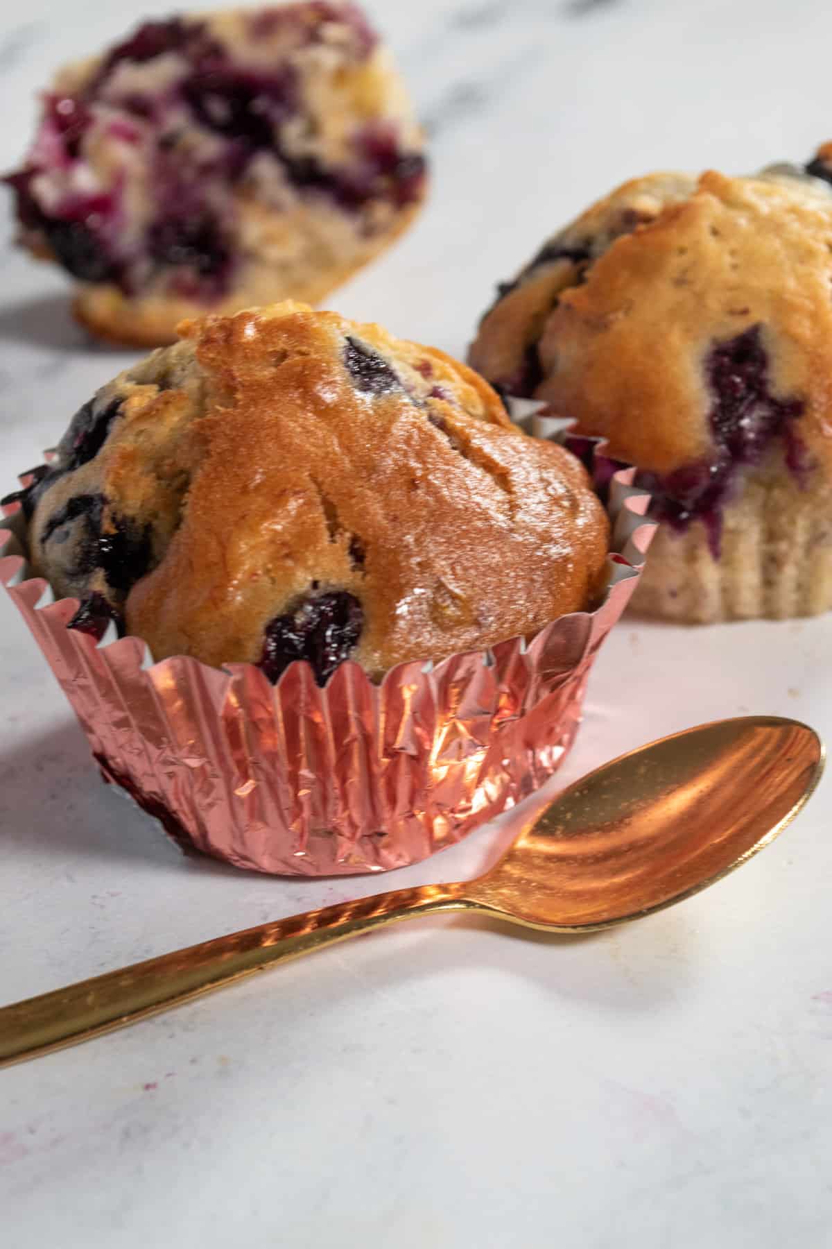 A close up shot of three pistachio muffins with blueberries looking juicy.  A golden spoon lies in front of them. 