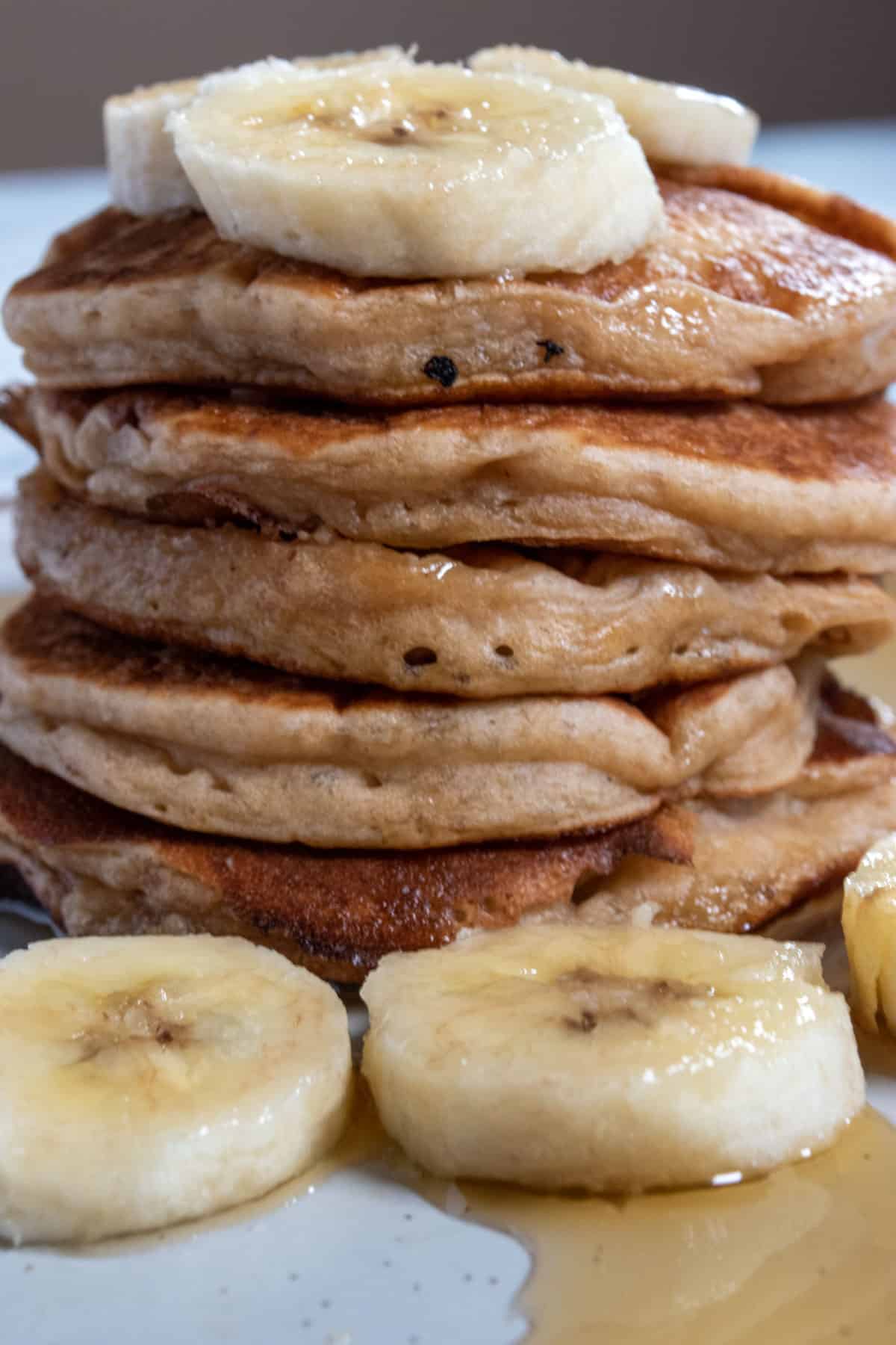 Golden brown and thick pancakes stacked into a tall pile. They are topped with chopped bananas. The are also bananas sat beside the base of the stack.