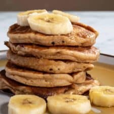 A stack of vegan banana protein pancakes. Decorated with maple syrup and chopped bananas.