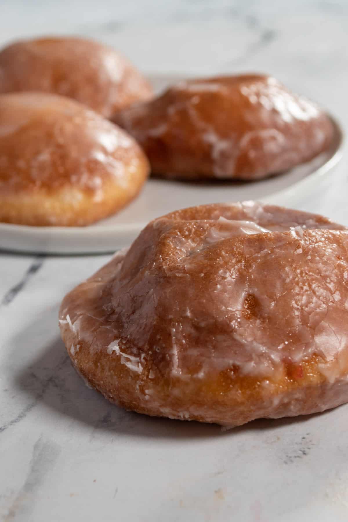 Four glazed donuts. One in the foreground and three on a plate in the background. 
