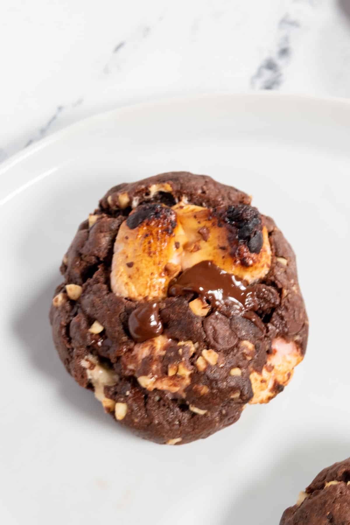 A shot of a round rocky road cookie. Two large marshmallows bursting out of the top. 