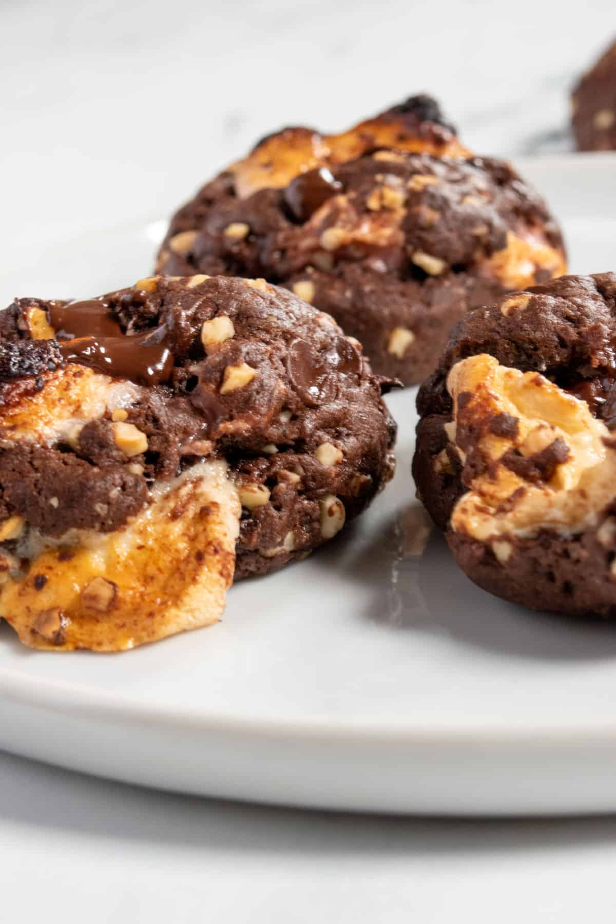 Three cookies §on that white plate. Marshmallow oozing from the cookie in the foreground. Melted chocolate on top. 