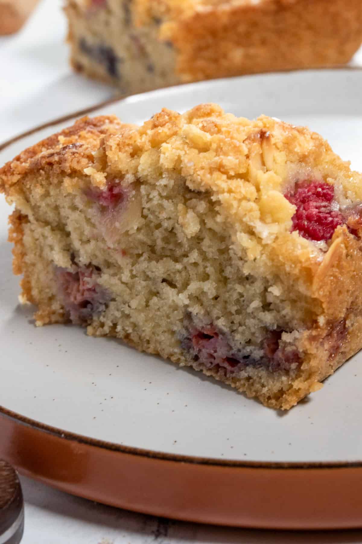 A single slice of almond raspberry cake on a brown-rimmed plate. There are lots of raspberries inside the cake, as well as on top. 