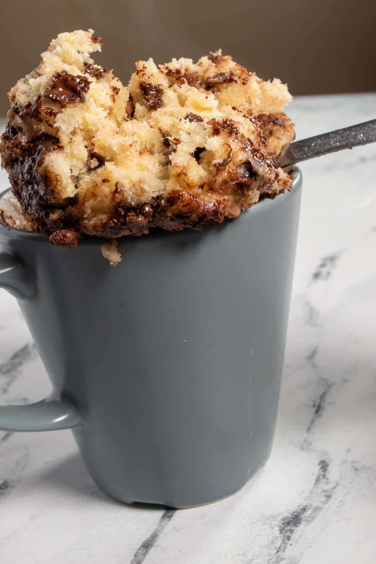 Cake being lifted out of a mug on a silver spoon. The vegan banana mug cake is hot and steam is rising upwards. 