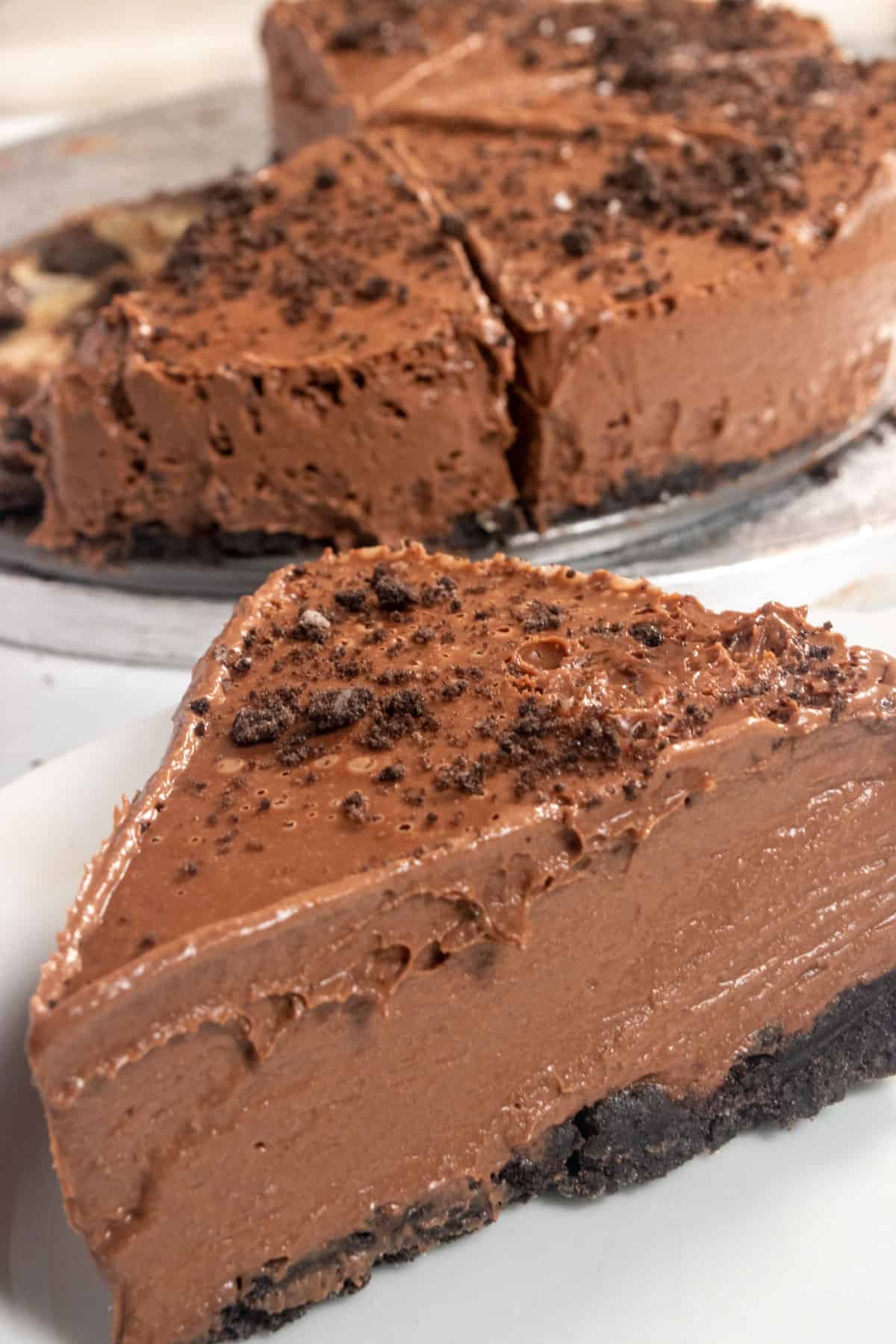 A chunky, chocolatey slice of my vegan chocolate cheese cake on a small white plate. Several more slices are on a platter in the background. 