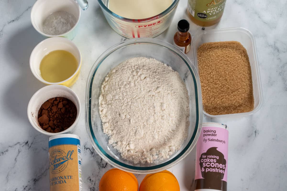 The ingredients needed to make a vegan chocolate orange cake, laid out neatly over a white, marble background.