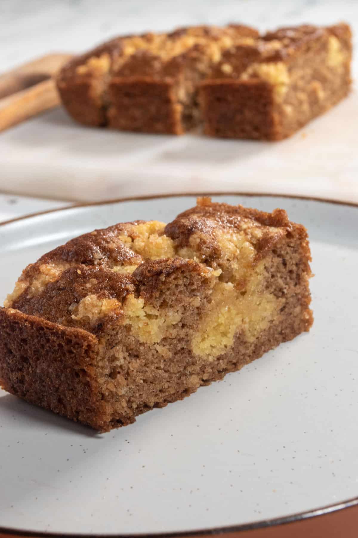 A small slice of vegan cinnamon cake on a brown-rimmed white plate. The rest of the loaf cake is on a marble platter in the background. 