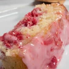 A slice of my vegan lemon raspberry cake on a white plate. It is topped with bright pink glaze and there are raspberries inside the cake.