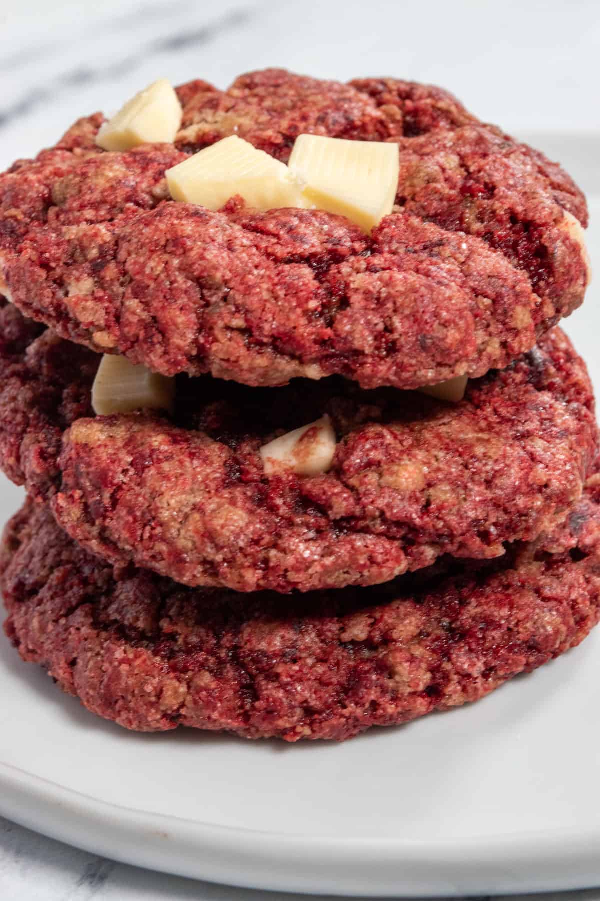 Three large vegan red velvet cookies stacked on a white plate. White chocolate is on top, melting.