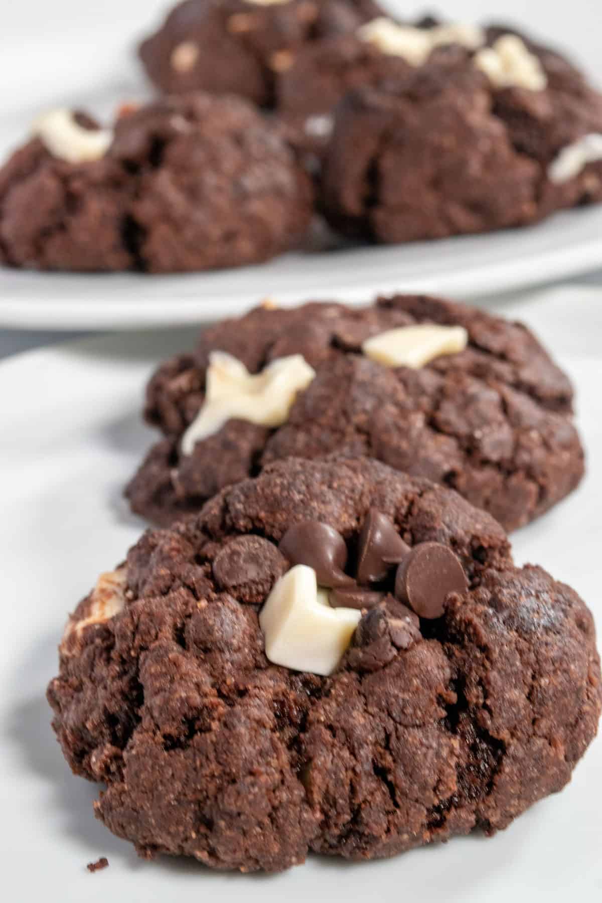 Two vegan triple chocolate cookies on a white plate. In the background, many more cookies are on a white platter.