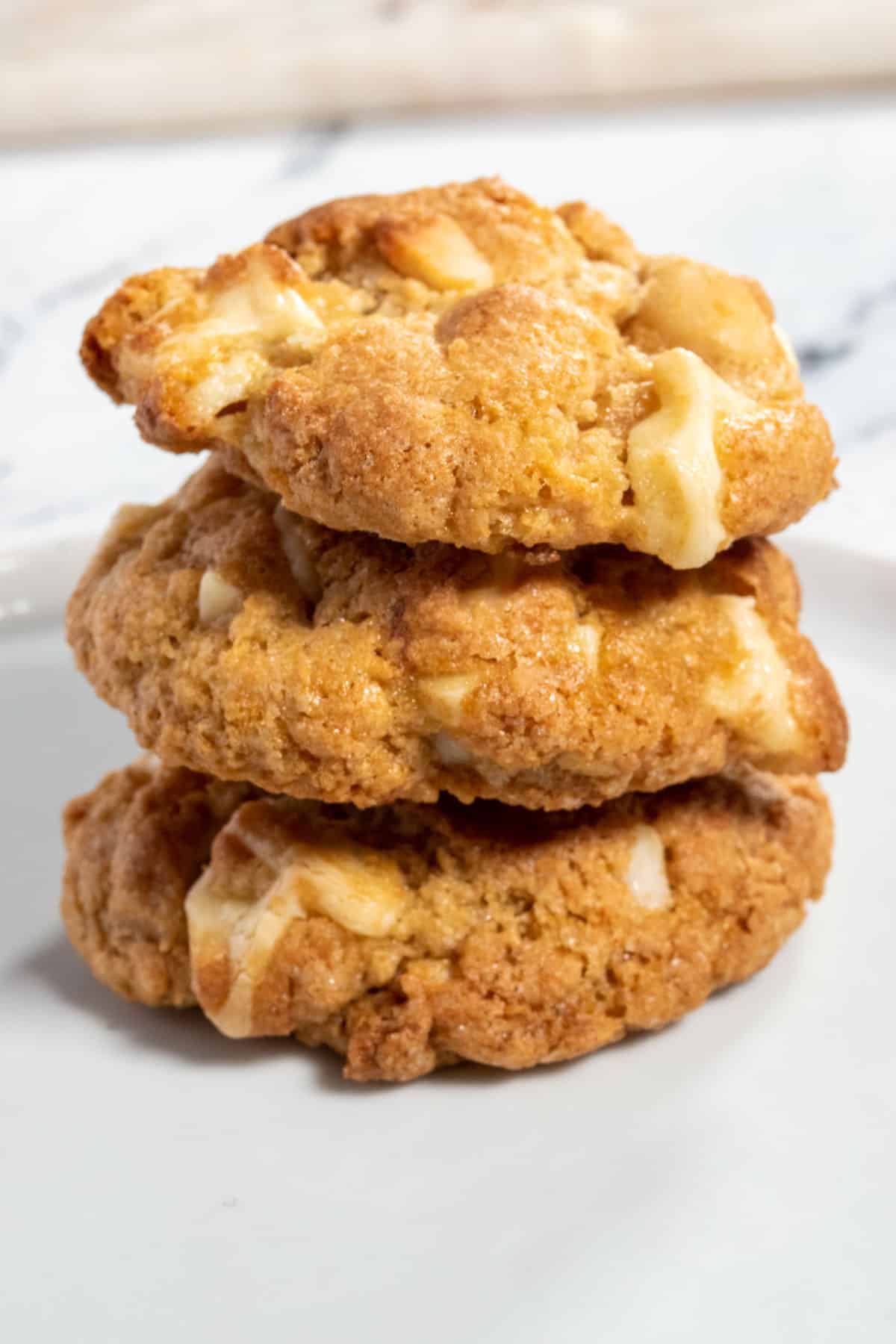 Three chunky vegan white chocolate cookies are stacked. They are very golden in colour. 