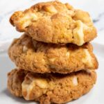 Three golden brown, vegan white chocolate cookies stacked in front of a white background.