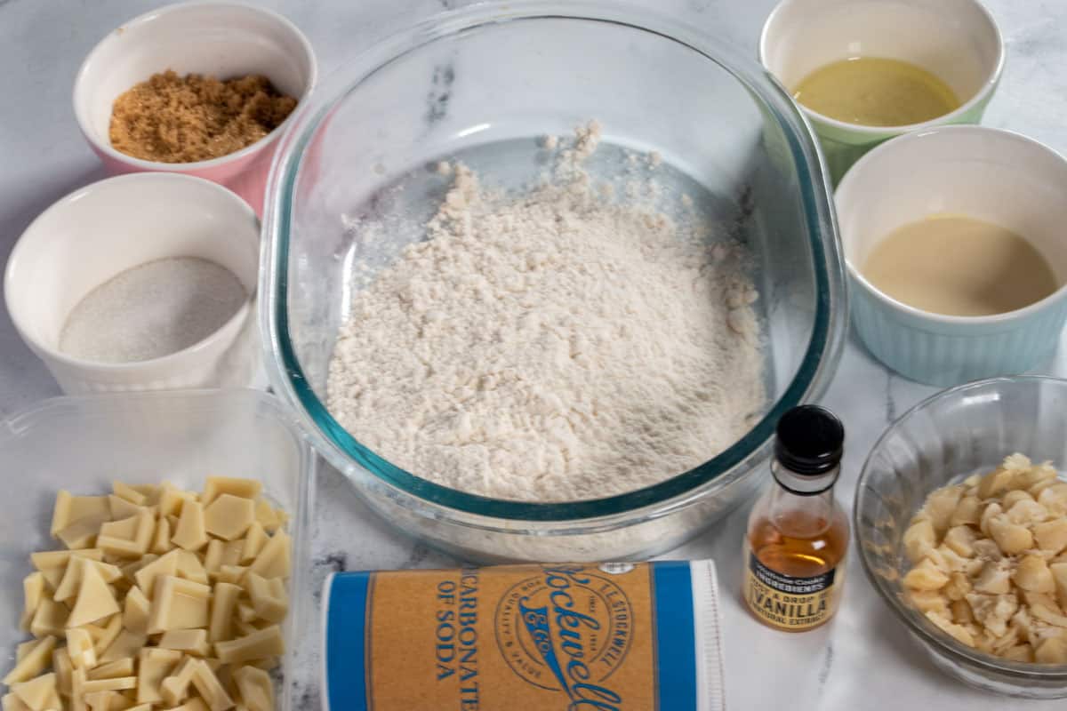 The ingredients needed to make vegan white chocolate macadamia nut cookies laid out over a white, marble background. 