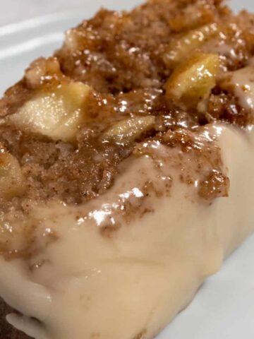 A thick slice of vegan apple cinnamon cake on a shiny, white plate. It is very moist and topped with maple glaze.