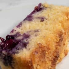 A thick slice of my vegan blueberry lemon cake on a shiny, white serving plate. The blueberries are bright purple inside the cake.