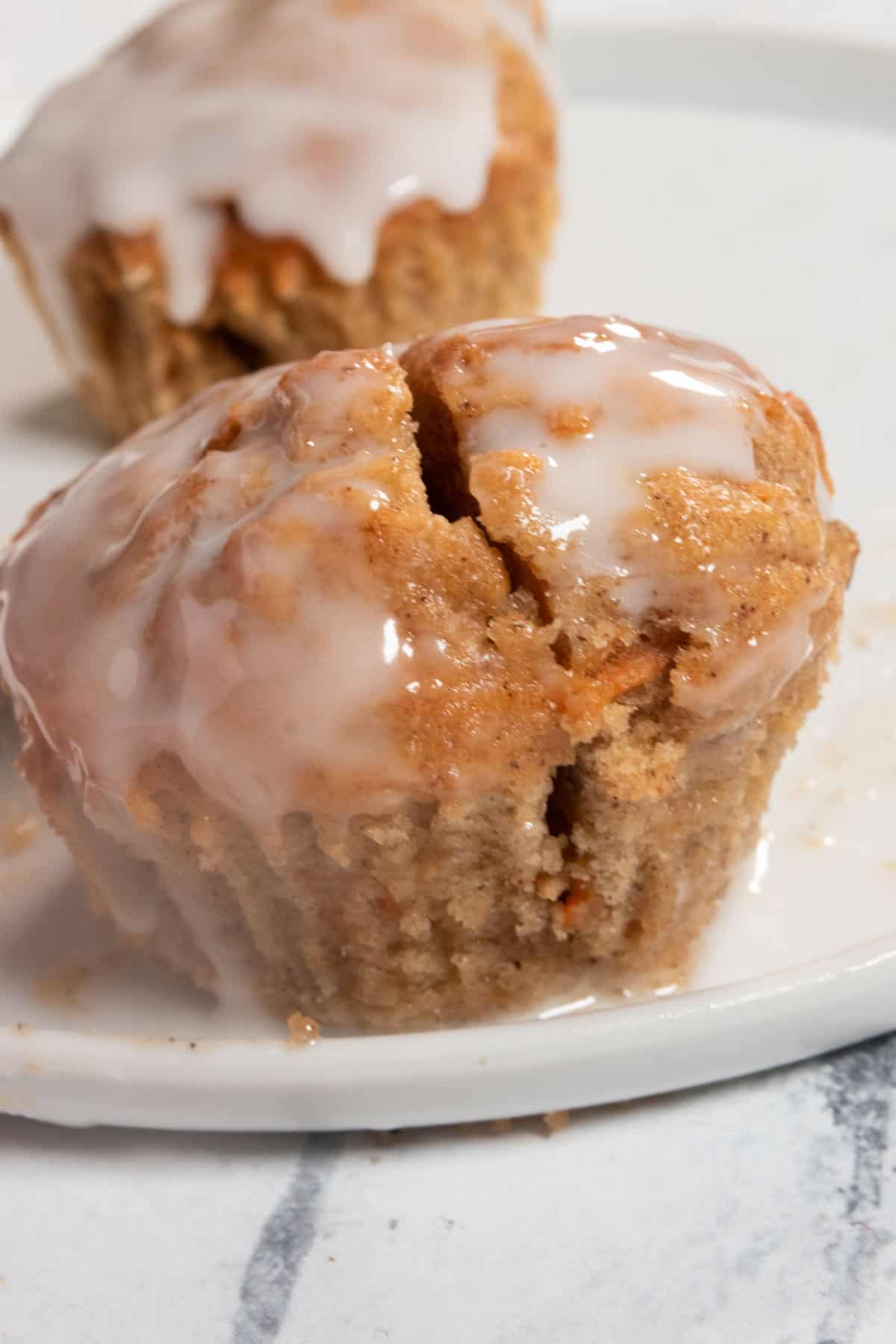 Two glazed vegan carrot cake muffins  on a shiny white plate. One is chopped in half. 