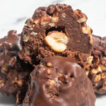 A small stack of my vegan ferrero rocher truffles on a white plate. One of them has been sliced open, revealing a hazelnut in the centre.