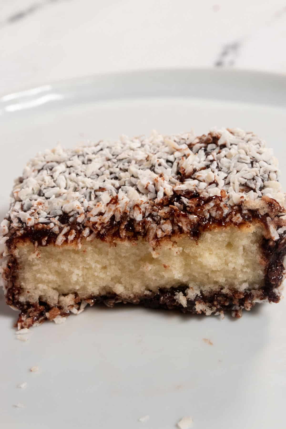A vegan Lamington bar on a white serving plate. The front of the cake has been sliced off revealing the soft cake in the center.
