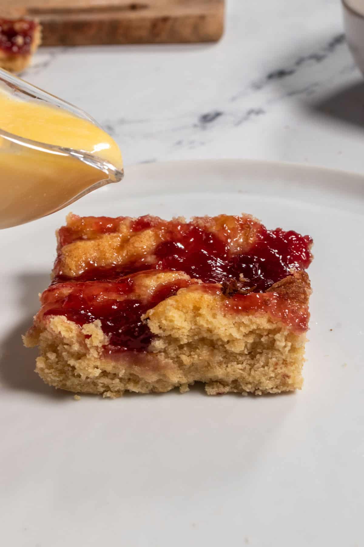 A small jug of vegan custard being poured over a slice of my vegan pudding cake with raspberry jam. 