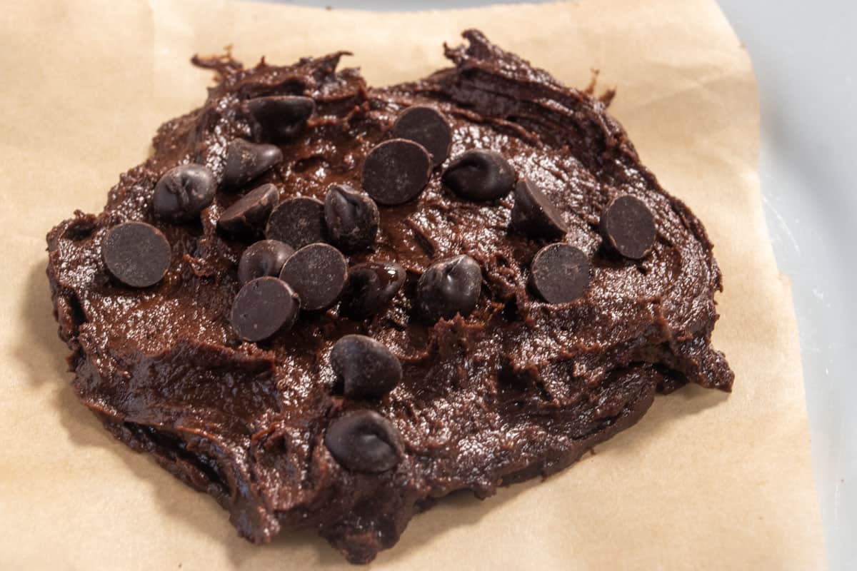 The chocolate cookie dough has been formed into a cookie shape on a piece of parchment paper. Chocolate chips have also be sprinkled on top. 