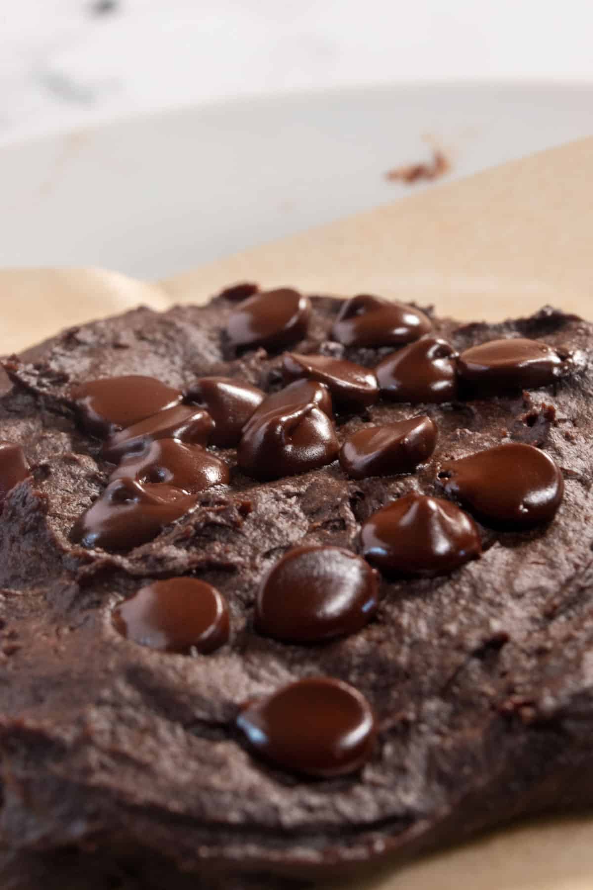 A baked single serve vegan cookie cooling on brown parchment paper. The chocolate chips on top are melty. 