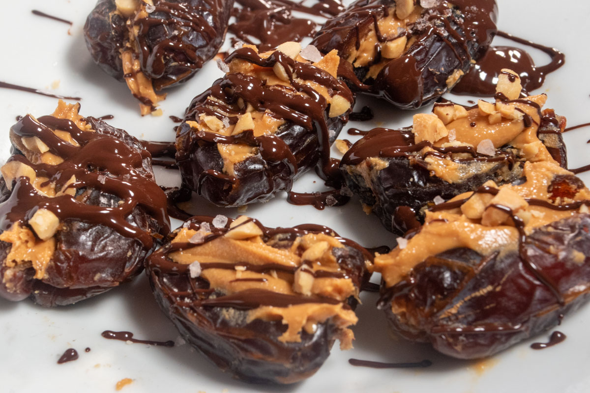 Lots of chocolate covered dates on a white platter. They're also topped with chopped peanuts, peanut butter and sea salt. 