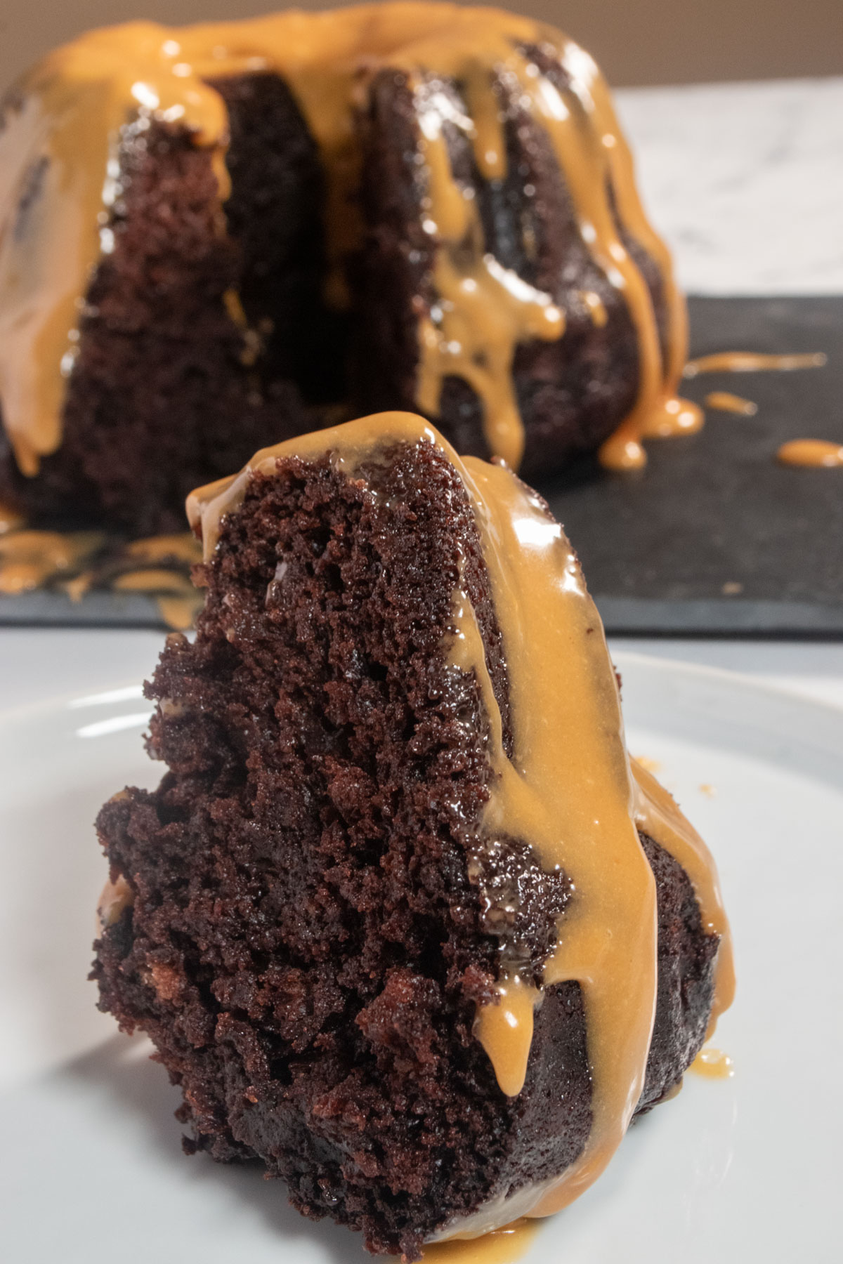 A huge slice of vegan chocolate bundt cake on a white saucer. The rest of the cake is sitting behind it on a slate platter, covered in peanut butter glaze. 