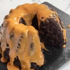 An almost-whole vegan chocolate bundt cake on a slate serving board covered in peanut butter glaze. One slice is missing.