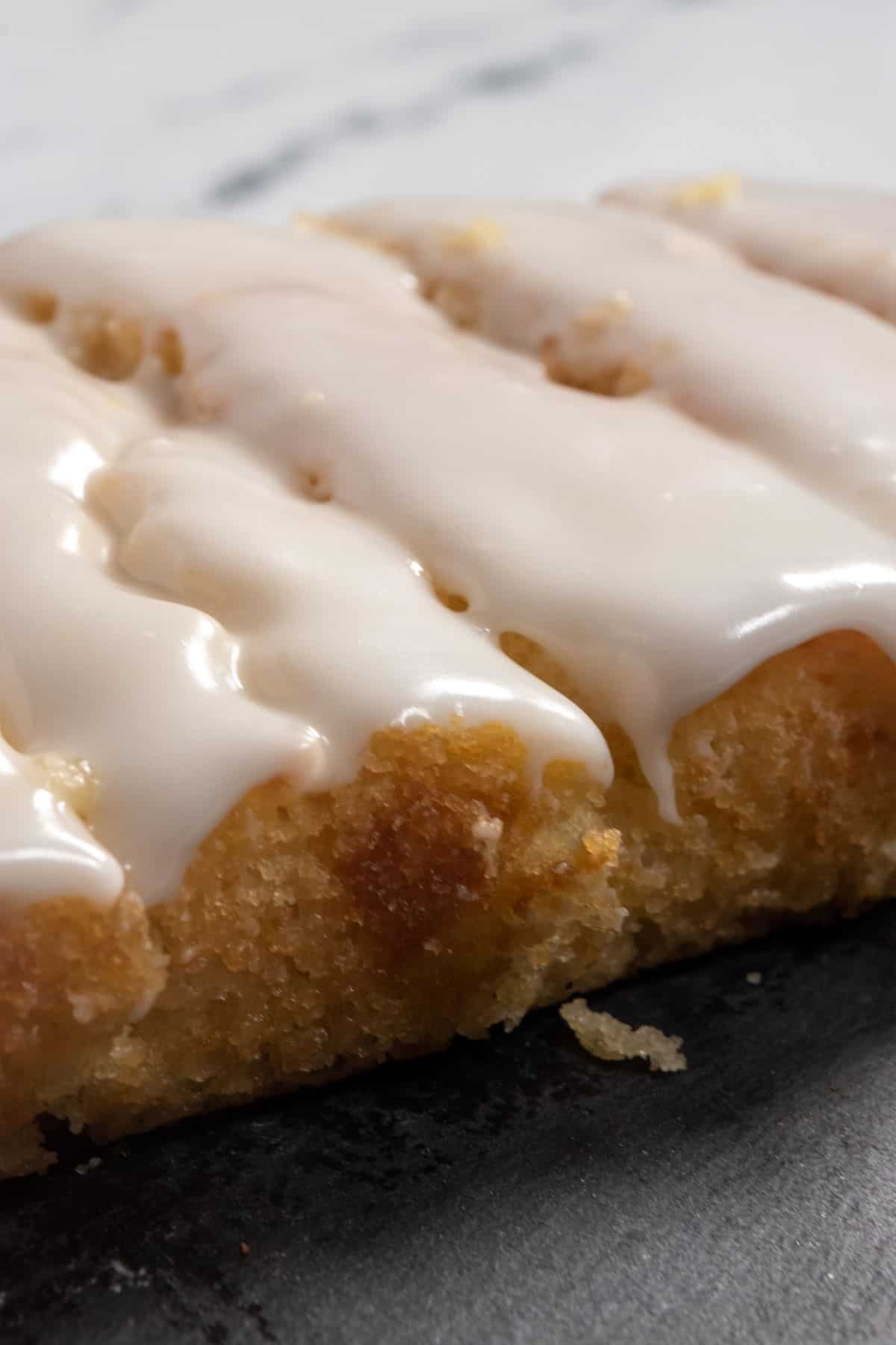 A whole lemon cake topped with melty glaze which is dripping down the sides. It is sat on a slate chopping board. 