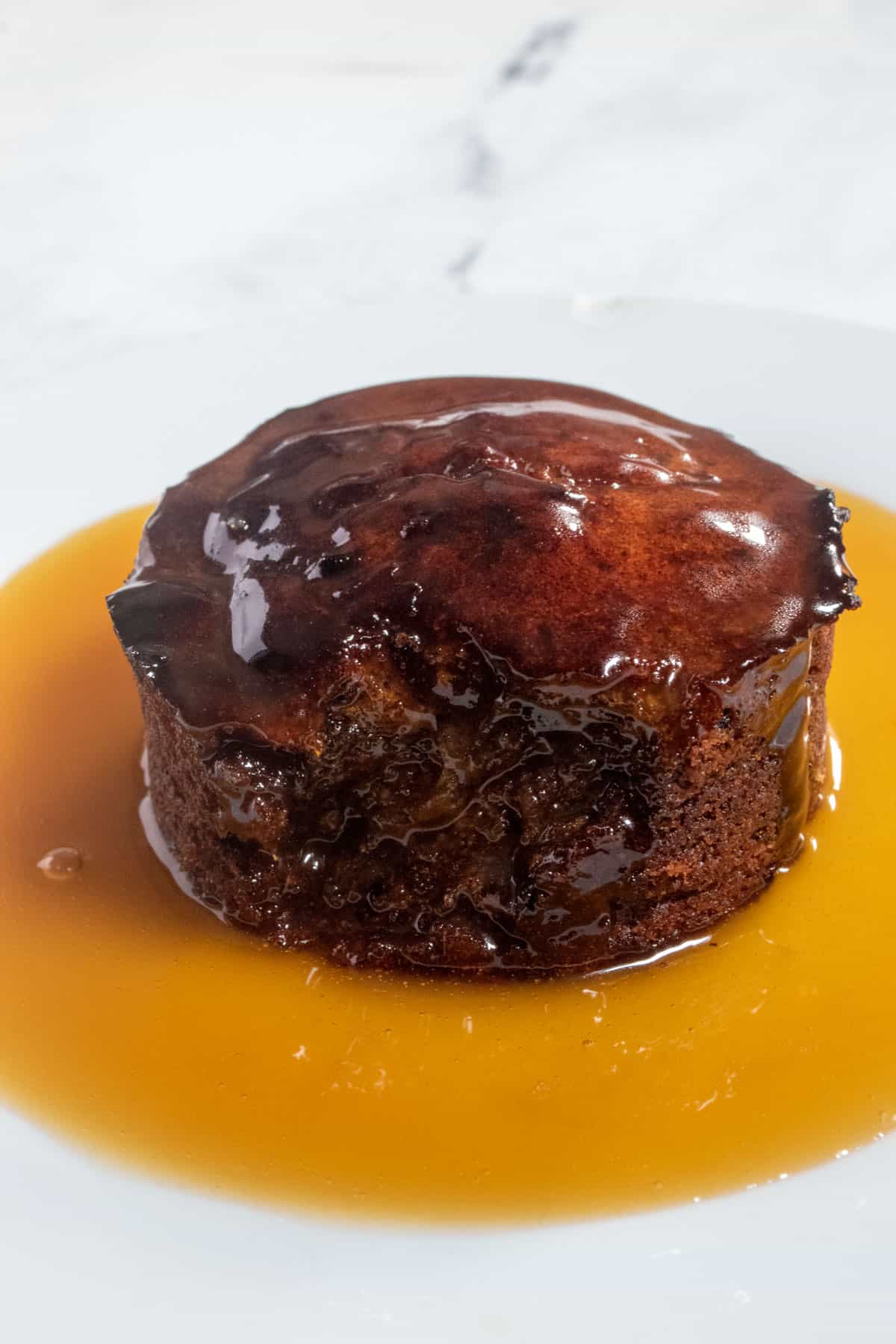 A whole vegan sticky toffee pudding cake on a white plate covered with vegan toffee sauce. 