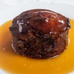 A whole vegan sticky toffee pudding covered in a pool of vegan toffee sauce.