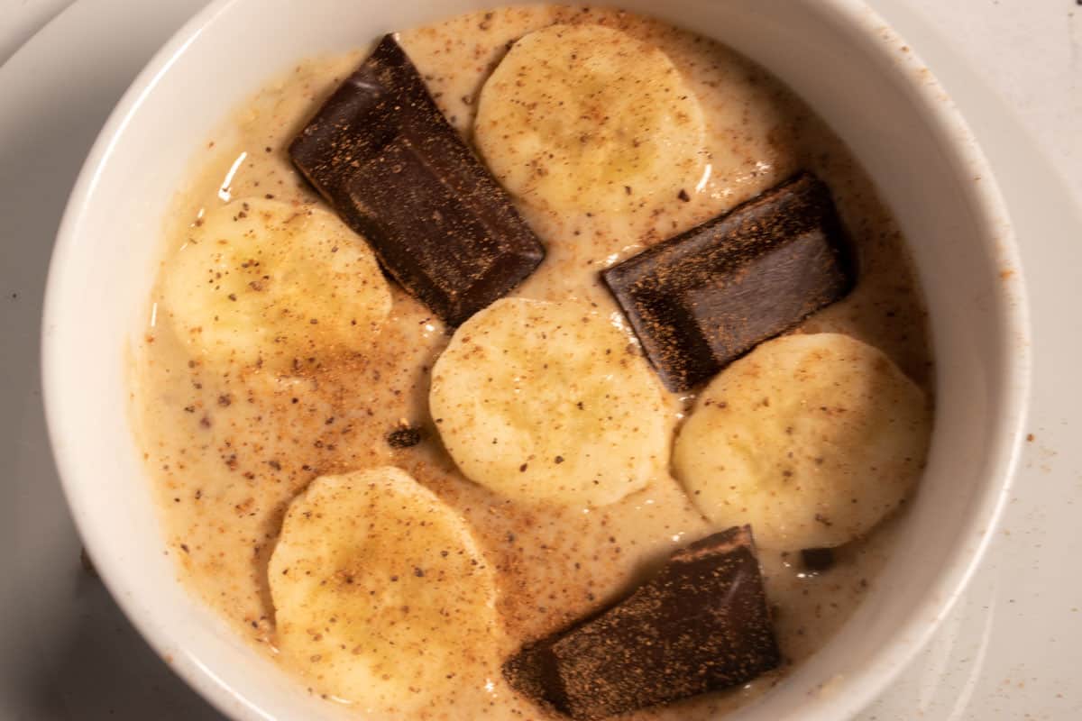 The banana oatmeal has been poured into a large ramekin. Chocolate chips and pecans have been mixed in it has been topped with chocolate chunks, cinnamon and banana slices. 