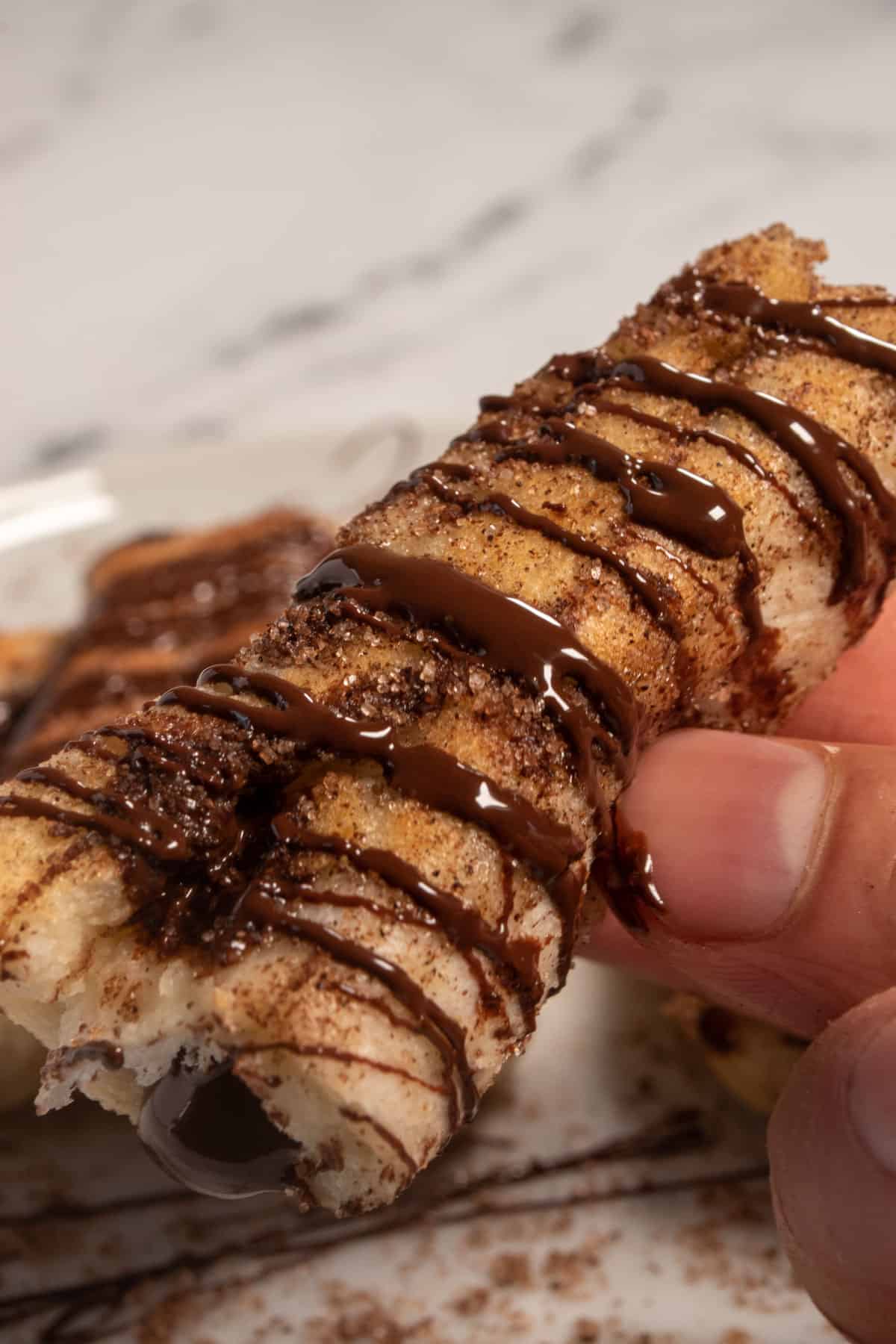 A chocolate french toast roll being held up by a hand. It is drizzled generously with chocolate. 