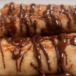 A close up image of stacked, golden brown chocolate french toast rolls drizzled with melted chocolate.