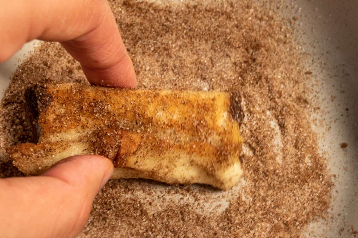 The french toast rolls being coated in cinnamon sugar by a hand.