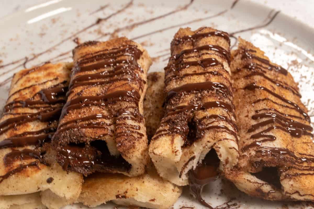 A stack of chocolate french toast rolls on a white plate. They are garnished with drizzled, melted vegan chocolate. 