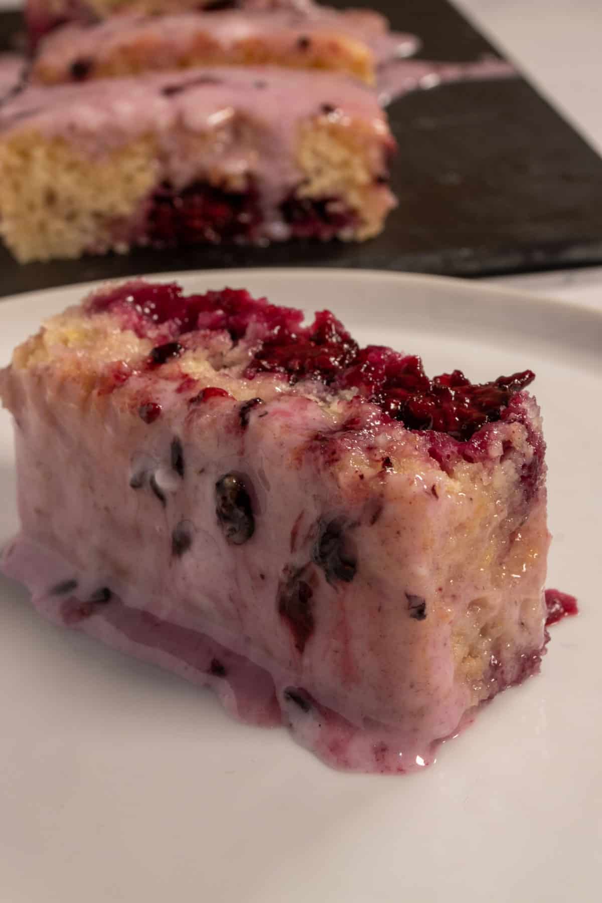 A juicy slice of vegan blueberry cake laying on its side on a white plate. The rest of the cake is in the background. 
