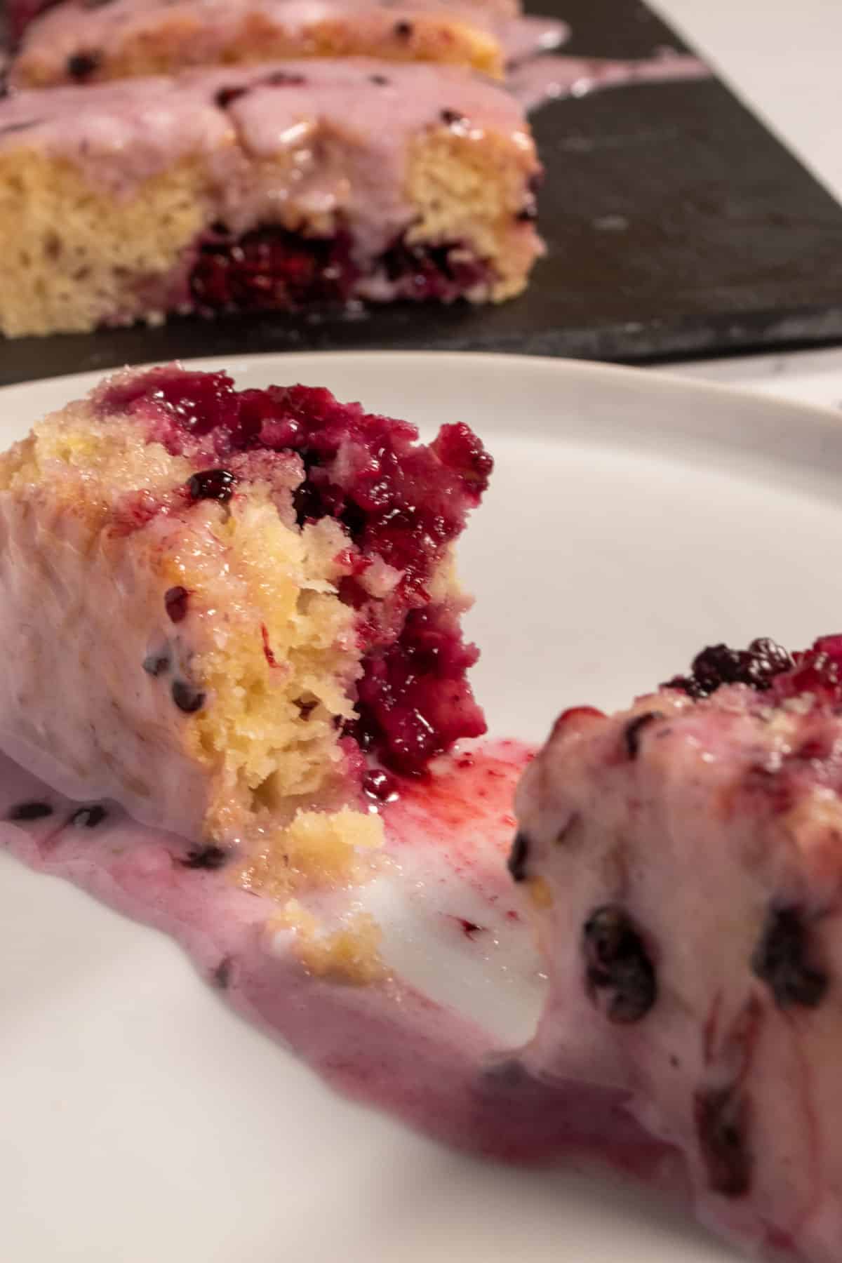 A piece of cake which has been sliced in half revealing lots of blackberries on the inside. It sits on a small, white plate, while the rest of the cake is in the background.