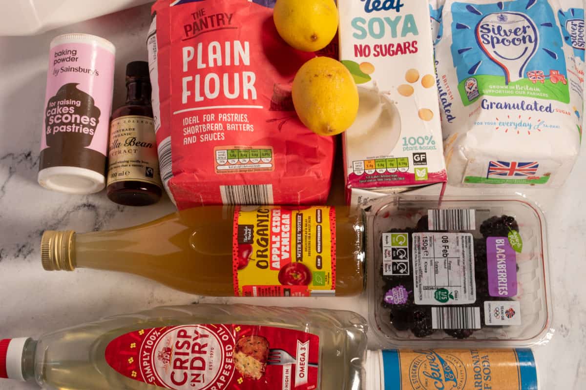 The ingredients that are needed to make a vegan blackberry cake laid out over a white, marble background. 